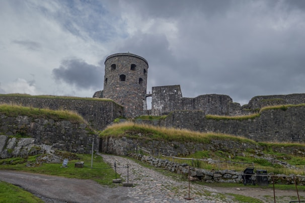 A stone fortress stands prominently against a cloudy sky, surrounded by lush green grass and low shrubs. The structure features a large cylindrical tower and stout, well-fortified walls made from dark stone. A cobblestone path leads to the entrance, adding to the historical atmosphere. The scene appears tranquil yet imposing.