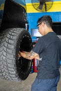 A man with glasses and a tattoo on his arm is working on a large vehicle's wheel. The setting appears to be a workshop with a blue and yellow wall in the background and a fan mounted on the wall above.