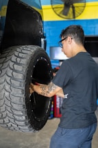 A man with glasses and a tattoo on his arm is working on a large vehicle's wheel. The setting appears to be a workshop with a blue and yellow wall in the background and a fan mounted on the wall above.