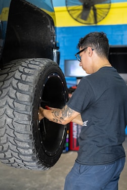 A man with glasses and a tattoo on his arm is working on a large vehicle's wheel. The setting appears to be a workshop with a blue and yellow wall in the background and a fan mounted on the wall above.