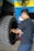 A man with glasses and a tattoo on his arm is working on a large vehicle's wheel. The setting appears to be a workshop with a blue and yellow wall in the background and a fan mounted on the wall above.