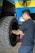A man with glasses and a tattoo on his arm is working on a large vehicle's wheel. The setting appears to be a workshop with a blue and yellow wall in the background and a fan mounted on the wall above.