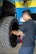 A man with glasses and a tattoo on his arm is working on a large vehicle's wheel. The setting appears to be a workshop with a blue and yellow wall in the background and a fan mounted on the wall above.