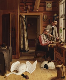 A warmly lit wooden room features two men, one sitting at a table and the other standing behind him, engaged in some form of craftwork. They are surrounded by various wooden tools, a clock on the wall, and clothing hanging from hooks. On the floor, a large white swan lies motionless near a small black-and-white dog curled up sleeping. The window lets in natural light, casting soft shadows across the room.