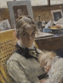 A woman with curly hair is seated in a wooden chair, wearing a formal, ruffled dress. She holds a sleeping child on her lap. The background features a cluttered desk with framed photographs, bottles, and various papers, suggesting a personal or artistic workspace.