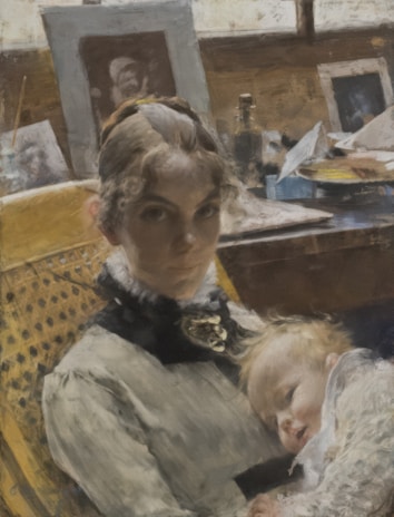 A woman with curly hair is seated in a wooden chair, wearing a formal, ruffled dress. She holds a sleeping child on her lap. The background features a cluttered desk with framed photographs, bottles, and various papers, suggesting a personal or artistic workspace.