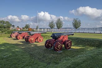 Several historic cannons are arranged on a grassy area in a row, with a backdrop of calm water and fluffy clouds in the sky. The cannons have red wooden carriages and are aimed outward towards the water. Trees and a few buildings can be seen in the background, contributing to a serene landscape.