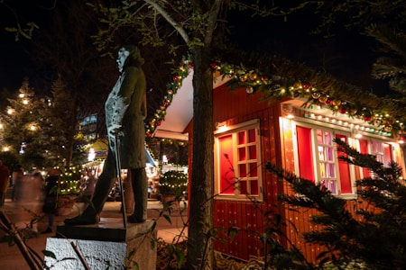 A festive outdoor scene featuring a bronze statue of a person standing on a pedestal beside a wooden cabin adorned with glowing Christmas lights and colorful ornament garlands. The cabin has red and white windows that enhance the holiday atmosphere. In the background, blurred people create a lively, bustling ambiance, with additional string lights illuminating the evening setting.