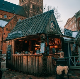A cozy wooden market stall adorned with string lights and seasonal decorations stands in front of a historic brick church. The sign reads 'Gingerbread House'. The stall offers various treats and is decorated with a star ornament. The background features snow on the rooftops, suggesting a winter setting.