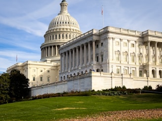 A grand neoclassical building with a large dome and pillars, set against a clear blue sky. The structure is surrounded by well-maintained lawns and a flag is visible on top of the building.