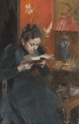A woman in a dark dress is deeply focused while reading a book. She is seated in a dimly lit room adorned with an ornate red wallpaper featuring floral patterns. In the background, a small table holds a decorative lamp with a red shade and a blue vase.