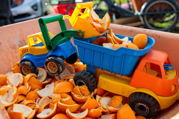 Toy construction vehicles, including a yellow and blue bulldozer and an orange and yellow dump truck, are filled with orange peels. The vehicles are placed inside a large container also containing several orange peels.