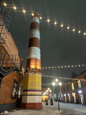 A tall, striped industrial chimney is illuminated against a dark evening sky, surrounded by strings of small lights. The chimney is situated in a brick-walled alley with buildings on either side that have brightly-lit windows. Snow covers the ground, adding a wintry atmosphere to the scene.