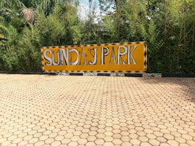 A hexagonal tiled pavement leads to a large yellow sign with bold black and white letters spelling 'SUNDAY PARK'. The sign is set against a backdrop of dense, green foliage, including bamboo and other plants, creating a vibrant natural setting.