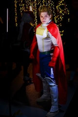 A person is dressed in a superhero costume featuring a red cape, white top with yellow and blue details, blue pants, and white boots. They are standing confidently in a dimly lit space with festive lights glowing in the background.