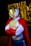 A person wearing a superhero costume with a white base, red gloves and cape, and a gold face mask. The background features blurred, twinkling lights adding a festive or theatrical atmosphere.