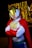 A person wearing a superhero costume with a white base, red gloves and cape, and a gold face mask. The background features blurred, twinkling lights adding a festive or theatrical atmosphere.