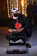A person dressed in a ninja-style outfit with a headband and mask is kneeling next to an open briefcase filled with stacks of hundred-dollar bills. The background features decorative string lights creating a festive atmosphere.