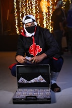 A person dressed in a ninja-style outfit with a headband and mask is kneeling next to an open briefcase filled with stacks of hundred-dollar bills. The background features decorative string lights creating a festive atmosphere.