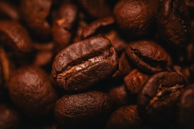 A close-up of roasted coffee beans with a rich, textured surface, highlighting their deep brown color and natural oil sheen. The beans are tightly clustered together, emphasizing their glossy appearance and robust texture.