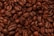 A close-up view of a heap of roasted coffee beans. The beans exhibit a rich, dark brown color and have a shiny, oily surface. Each bean is distinct, with visible textures and creases characteristic of roasted coffee beans.