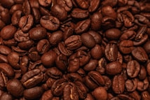 A close-up view of a heap of roasted coffee beans. The beans exhibit a rich, dark brown color and have a shiny, oily surface. Each bean is distinct, with visible textures and creases characteristic of roasted coffee beans.