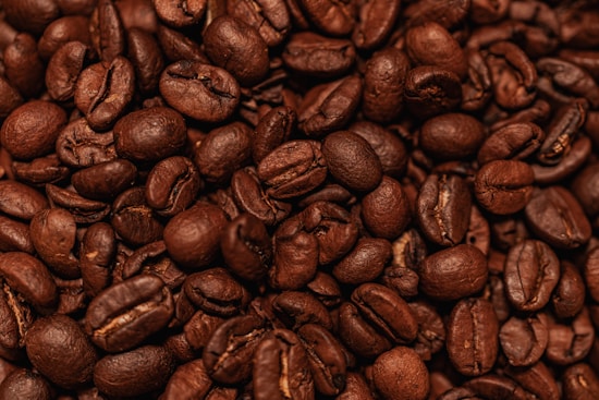 A close-up view of a heap of roasted coffee beans. The beans exhibit a rich, dark brown color and have a shiny, oily surface. Each bean is distinct, with visible textures and creases characteristic of roasted coffee beans.