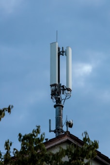A cell tower stands atop a building with a clear sky in the background. The tower features multiple antennas and cables, indicating its use for telecommunications. Partial foliage from nearby trees can be seen in the foreground.