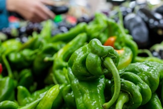 A close-up view of a pile of fresh green peppers with a hand holding a black object in the background. The peppers are shiny and vary slightly in size and shape. They are piled on a display at what appears to be a market or store setting.