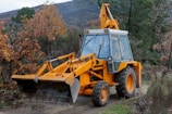 A yellow excavator with a front bucket stands on a dirt path surrounded by dense foliage and autumn-colored leaves. The machine appears ready for use, with visible hydraulic arms and large tires. The background features trees and a hillside under a cloudy sky.