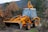A yellow excavator with a front bucket stands on a dirt path surrounded by dense foliage and autumn-colored leaves. The machine appears ready for use, with visible hydraulic arms and large tires. The background features trees and a hillside under a cloudy sky.