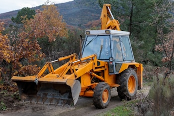 A yellow excavator with a front bucket stands on a dirt path surrounded by dense foliage and autumn-colored leaves. The machine appears ready for use, with visible hydraulic arms and large tires. The background features trees and a hillside under a cloudy sky.