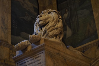 A majestic stone lion sculpture resting atop a pedestal. The lion exudes a sense of pride and strength with its detailed mane and serene expression. Surrounding the sculpture is an elegantly adorned interior with painted murals depicting historical scenes.