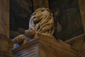 A majestic stone lion sculpture resting atop a pedestal. The lion exudes a sense of pride and strength with its detailed mane and serene expression. Surrounding the sculpture is an elegantly adorned interior with painted murals depicting historical scenes.