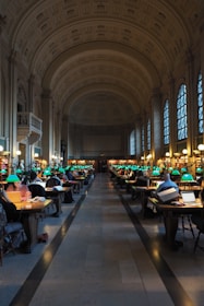 A grand library room with high vaulted ceilings and arched windows. Numerous people are seated at wooden tables, working on laptops and reading books. Each table is equipped with green-shaded lamps and the room has a central aisle leading to a distant wall filled with bookshelves.