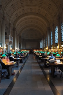A grand library room with high vaulted ceilings and arched windows. Numerous people are seated at wooden tables, working on laptops and reading books. Each table is equipped with green-shaded lamps and the room has a central aisle leading to a distant wall filled with bookshelves.