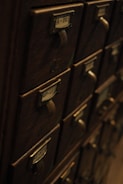 A collection of vintage wooden drawers with metal handles and label slots. The drawers have labels with various letters and numbers, giving the appearance of an old library catalog card system. The atmosphere is dimly lit, highlighting the aged wood texture.