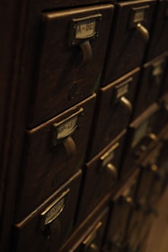 A collection of vintage wooden drawers with metal handles and label slots. The drawers have labels with various letters and numbers, giving the appearance of an old library catalog card system. The atmosphere is dimly lit, highlighting the aged wood texture.