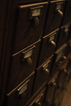 A collection of vintage wooden drawers with metal handles and label slots. The drawers have labels with various letters and numbers, giving the appearance of an old library catalog card system. The atmosphere is dimly lit, highlighting the aged wood texture.