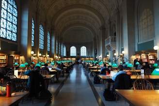 A grand library hall with tall arched windows and elaborate moldings on the ceiling. Rows of large wooden tables are lined with green reading lamps, and people are seated, engaged in reading or using laptops. The shelves are filled with numerous books, and the ambiance is quiet and studious.