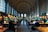 A grand library hall with tall arched windows and elaborate moldings on the ceiling. Rows of large wooden tables are lined with green reading lamps, and people are seated, engaged in reading or using laptops. The shelves are filled with numerous books, and the ambiance is quiet and studious.