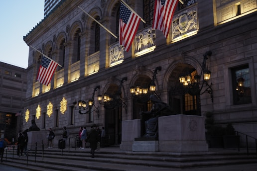 A grand stone building with intricate carvings and large arched windows is adorned with illuminated holiday decorations. Three American flags are prominently displayed above the entrance, which is lined with ornate street lamps. People are gathered on the steps leading to the entrance, and a statue is visible next to the steps.