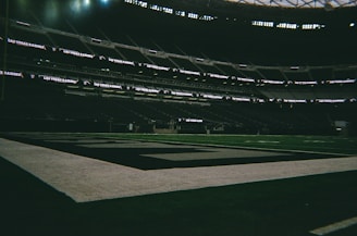 A dimly lit, empty football stadium with Las Vegas Raiders branding visible on the digital displays. The field appears well-maintained, with clear markings and a large goal area.