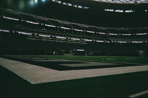 A dimly lit, empty football stadium with Las Vegas Raiders branding visible on the digital displays. The field appears well-maintained, with clear markings and a large goal area.