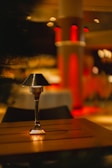 A small table lamp with a metal shade stands on a wooden table in a dimly-lit environment with a warm and cozy ambiance. The background features soft focus lighting and red-colored pillars, adding to the intimate atmosphere.