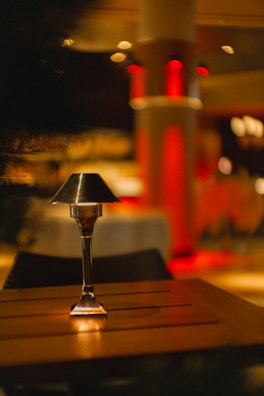 A small table lamp with a metal shade stands on a wooden table in a dimly-lit environment with a warm and cozy ambiance. The background features soft focus lighting and red-colored pillars, adding to the intimate atmosphere.