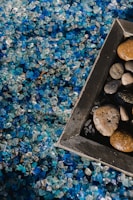 Close-up view of a mixture of small, decorative blue and clear stones, with a wooden-framed area containing a variety of round, smooth pebbles. The image captures the contrast between the colorful crystals and the muted earth tones of the pebbles.