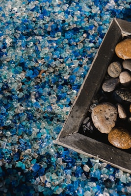 Close-up view of a mixture of small, decorative blue and clear stones, with a wooden-framed area containing a variety of round, smooth pebbles. The image captures the contrast between the colorful crystals and the muted earth tones of the pebbles.