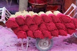 Stacks of incense sticks with vibrant red dyed tops are arranged on a wooden cart. The background features various red and pink colored containers and surfaces, suggesting a setting involved in the production or sale of incense.