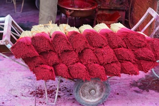 Stacks of incense sticks with vibrant red dyed tops are arranged on a wooden cart. The background features various red and pink colored containers and surfaces, suggesting a setting involved in the production or sale of incense.